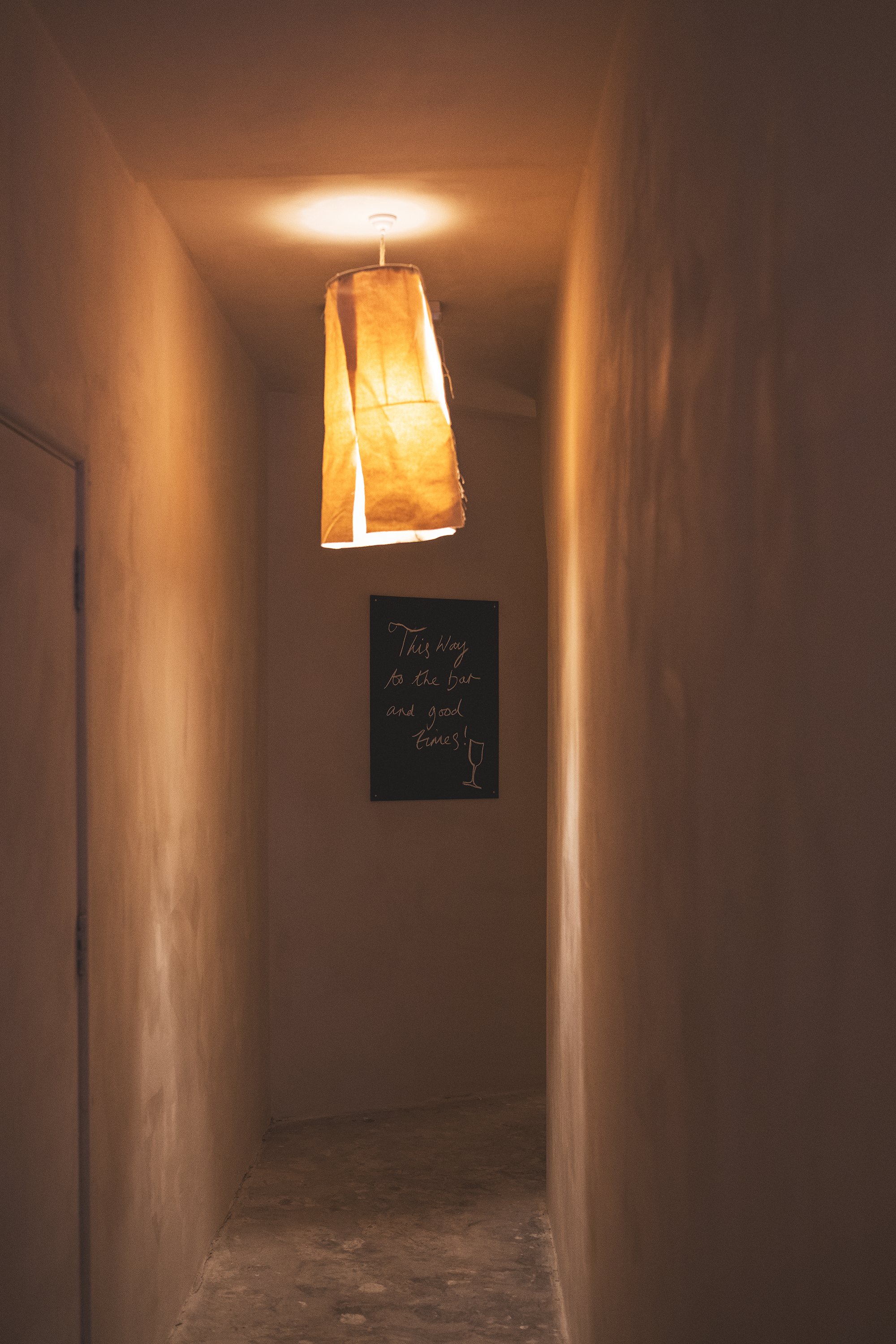 A dimly lit hallway with bare beige walls and a wooden floor. There is a hanging lamp with a fabric shade emitting warm light. A small black sign on the wall reads: "This way to the bar and good times!" with a wine glass illustration.