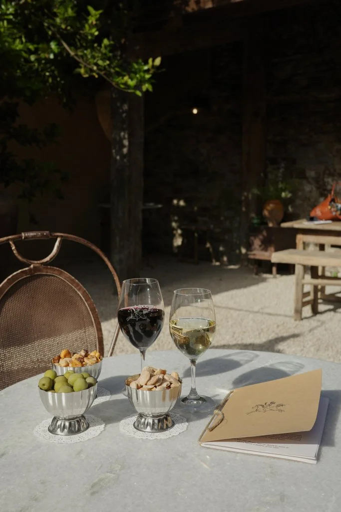 A table set outdoors with two glasses of wine, bowls of green olives, mixed nuts, and sliced chips, accompanied by a menu or brochure, with a rustic background and partial shade.