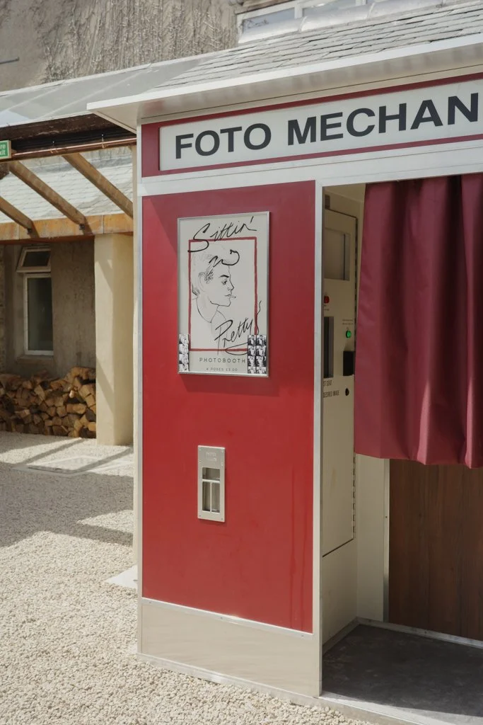 Photo booth kiosk with a sign that says "Foto Mechanic" and a poster of a person's outline with the handwritten phrase "Sittin' Pretty".