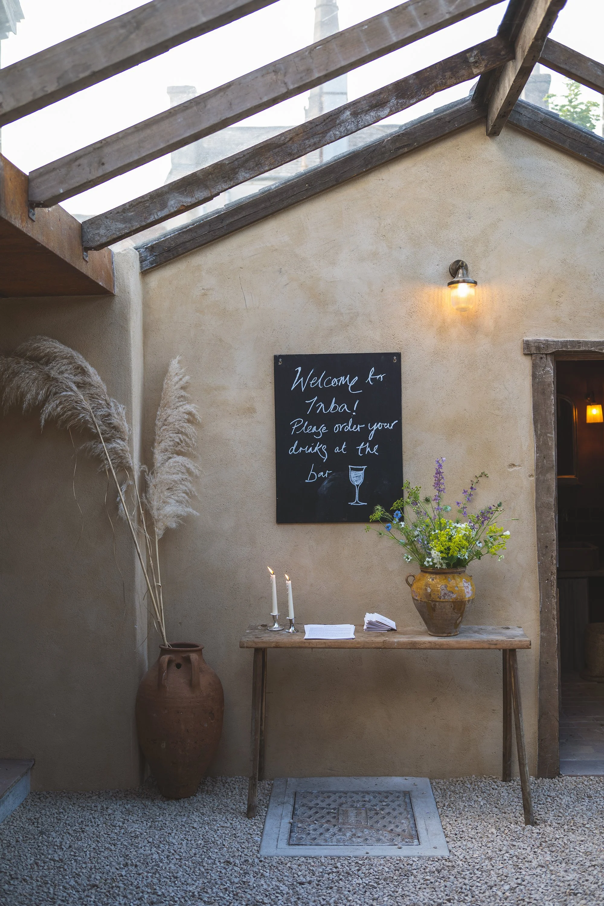A rustic outdoor courtyard area with a beige stucco wall, a wooden table with candles, a vase of colorful flowers, a chalkboard sign welcoming guests to Inaba, and a large pottery jar. There is a ceiling with wooden beams and a small alt light fixture.