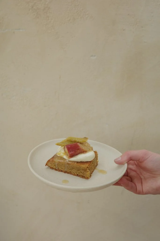 A person's hand holding a white plate with a slice of cake topped with whipped cream, a red fruit, and a yellowish garnish against a plain beige wall.