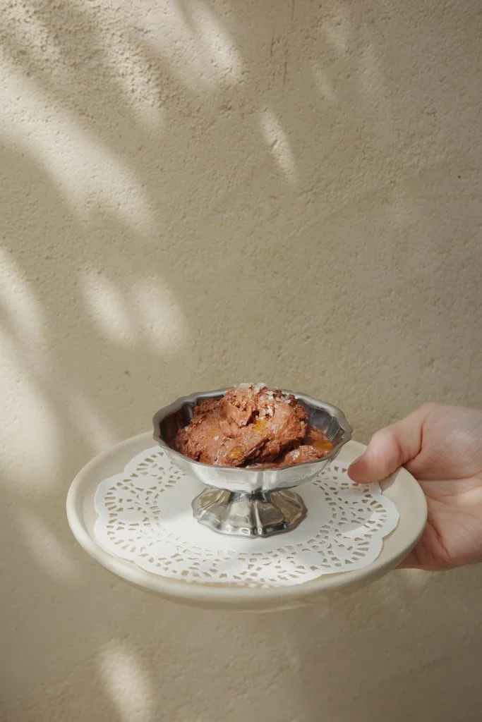 Hand holding a silver dish with a serving of chocolate chip cookie dough ice cream on a decorative paper doily, against a beige wall with soft shadows.