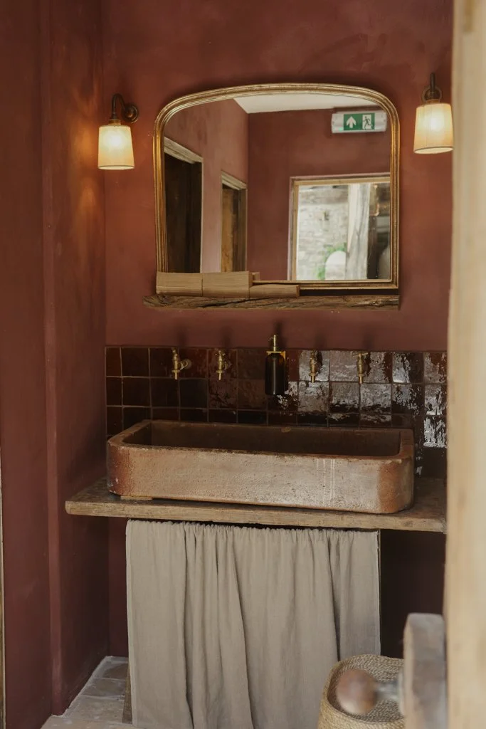 A rustic bathroom sink with a large stone basin, brass faucets, a mirror with a wooden frame, and warm lighting fixtures.