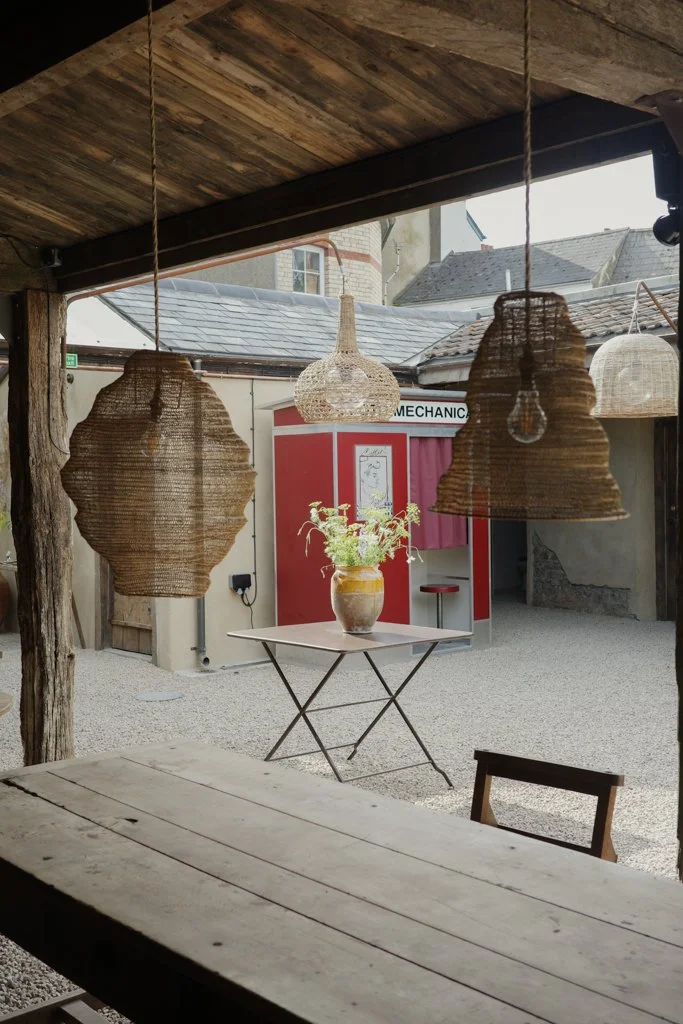Outdoor patio with a rustic wooden table, hanging woven lampshades, and a small table with a plant in a vase. In the background, there's a red booth marked "MECHANIC" and a bar stool.