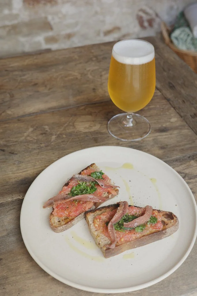 Two slices of pizza with anchovies, basil, and tomato on a white plate and a glass of beer on a wooden table.