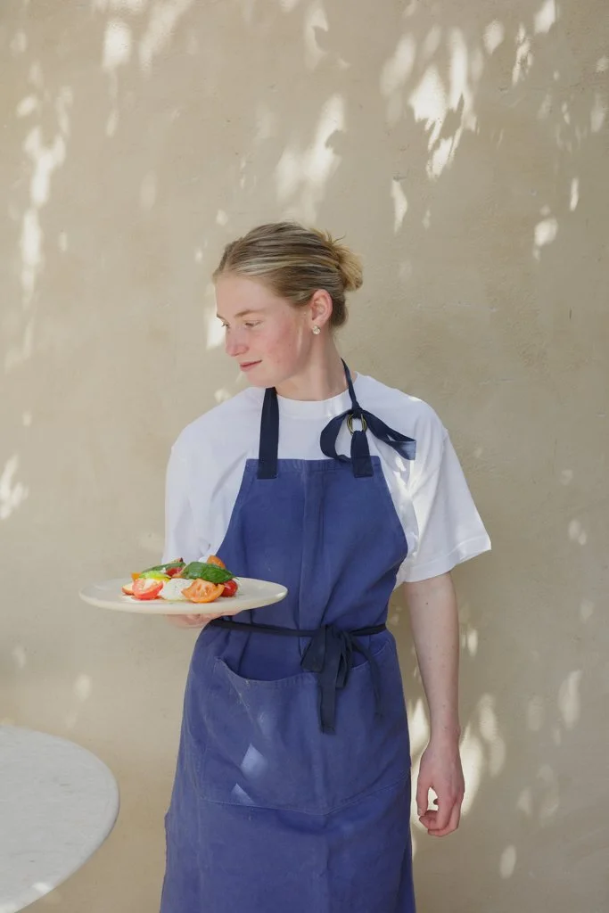A woman wearing a white shirt and blue apron holds a plate with an arrangement of fresh vegetables.