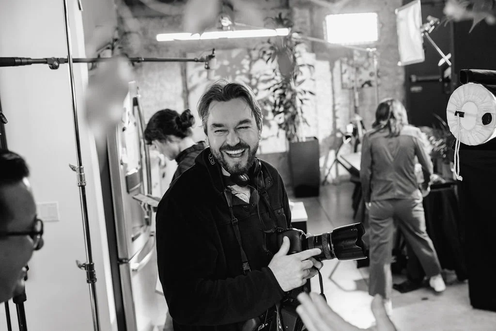 Photographer smiling and holding a camera in a studio with other people working in the background.