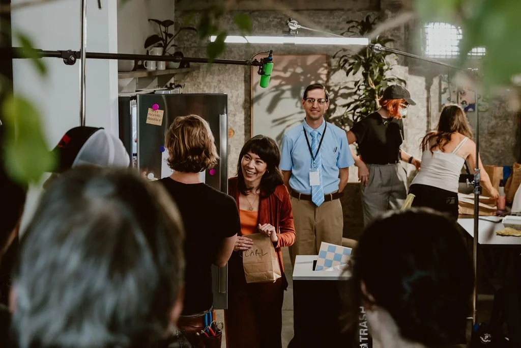 A group of people at an indoor event, some engaging in conversation near a table with a brown paper bag and decorative items, with additional people and activity in the background, amidst a modern, well-lit space with plants and commercial appliances