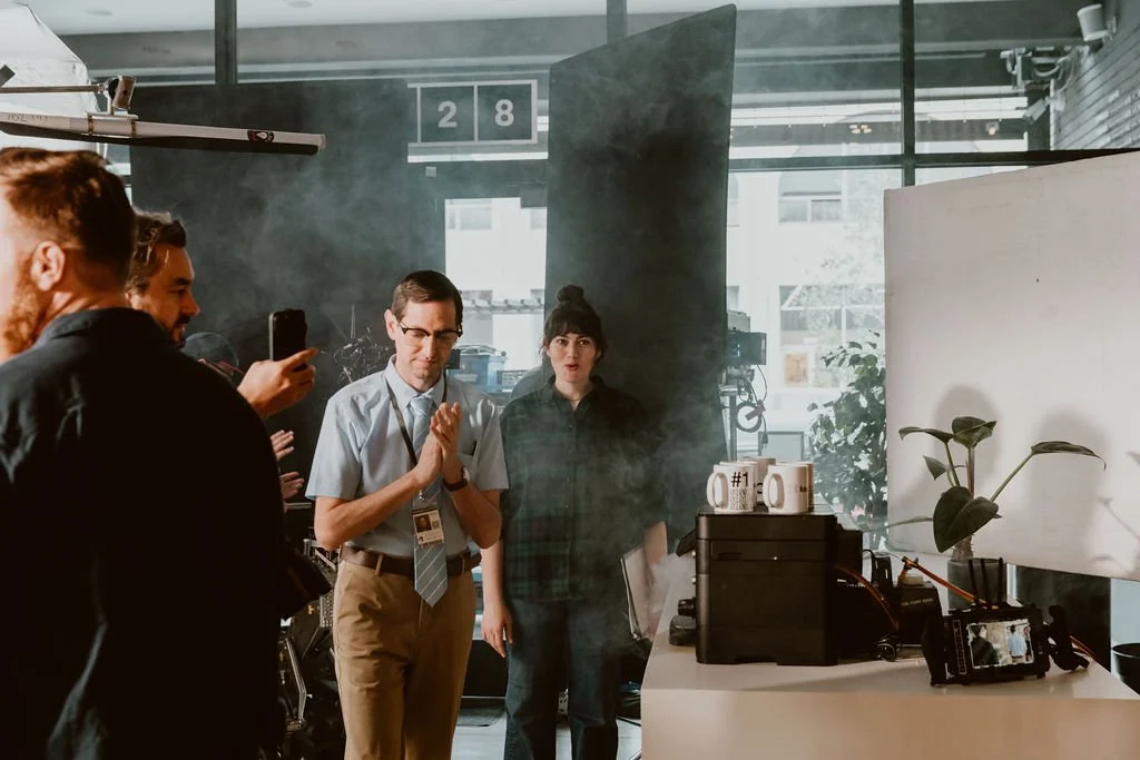 People in a photography studio with equipment, plants, and a white backdrop.