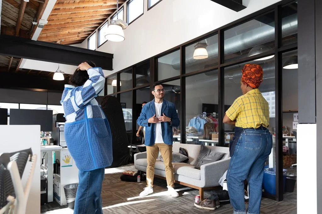 Three people engaged in conversation inside an office with large glass windows and a high ceiling.