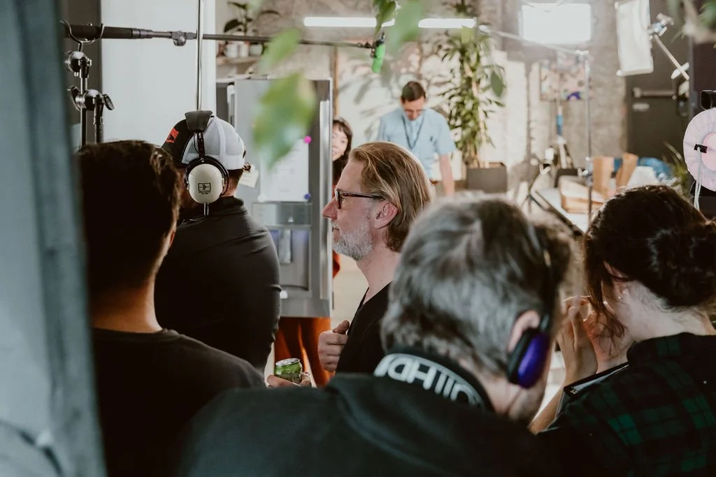 A group of people on a film or video set, with some wearing headphones and other equipment, in a room with various lighting and production gear, and a person in the background working on a desk.