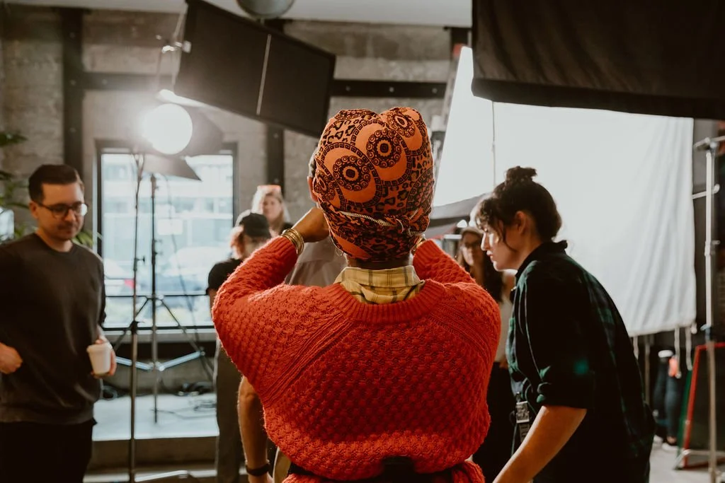 Behind-the-scenes moment on a film set with a woman in a colorful headscarf and red sweater, surrounded by crew members and equipment.