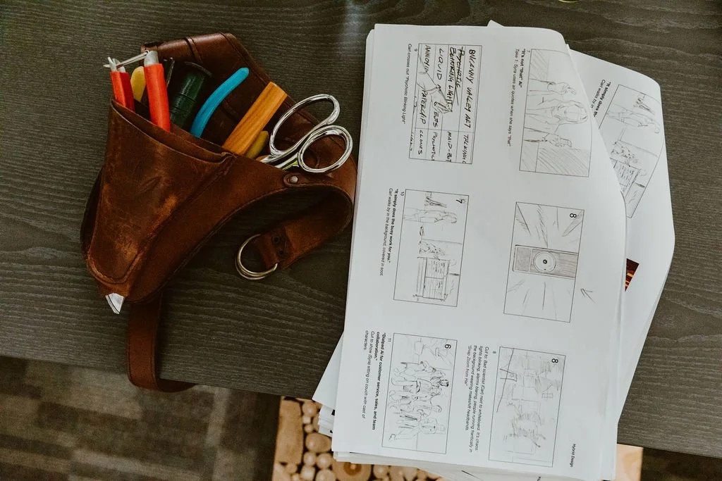 A leather pouch filled with pens, markers, and scissors beside scattered comic book or storyboard pages on a dark wooden table.