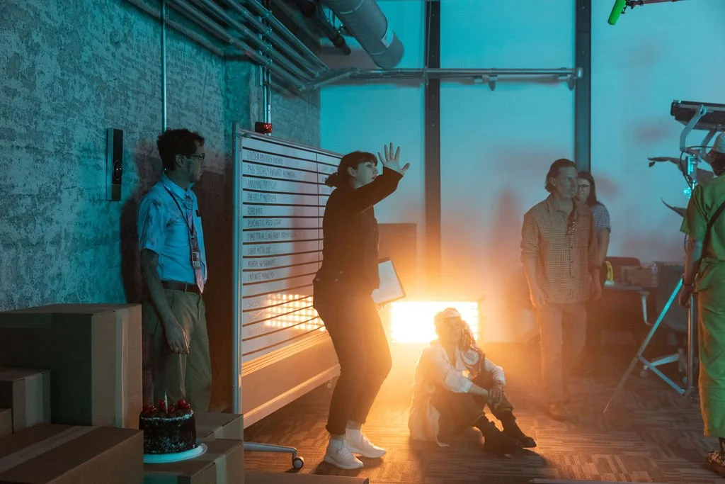 Group of people in a room with industrial decor, some standing and one sitting on the floor, with a bright light and equipment, possibly set for filming or photography.