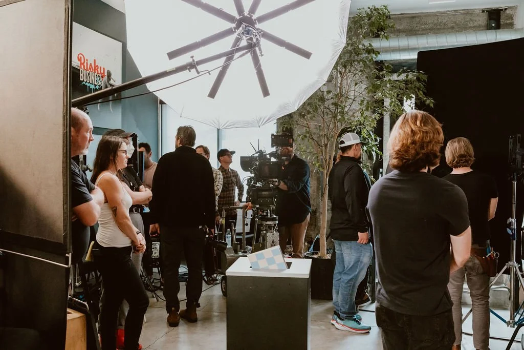 A group of people, including crew members and actors, are gathered in a studio set for a filming or photo shoot, with professional lighting equipment and cameras, and a sign reading 'Risky Business' in the background.