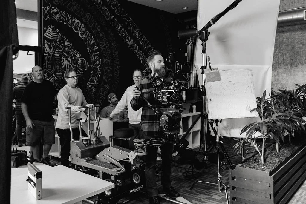 Film crew setting up equipment on a movie set, with a camera operator, lighting, and a white backdrop, and plants on the side.