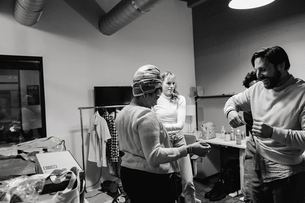 Three people in a room with clothing and supplies, one person wearing a headscarf, another woman in a white top, and a man smiling and adjusting his shirt.