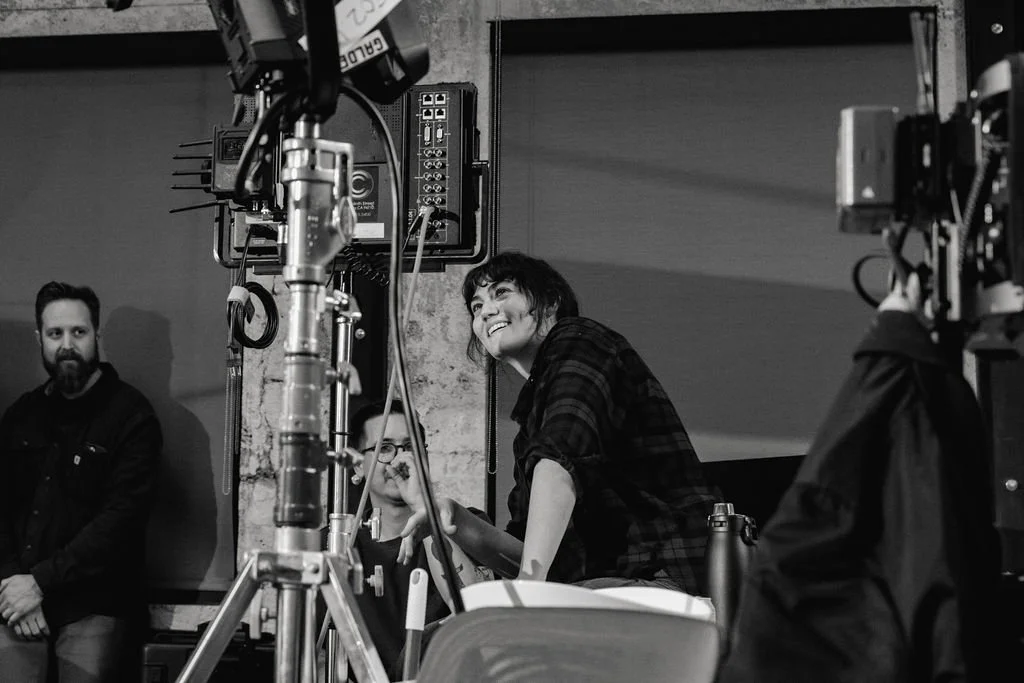 Black and white photo of a film set with a woman in a plaid shirt smiling and leaning on a surface, two people behind her, and crew members and equipment around.