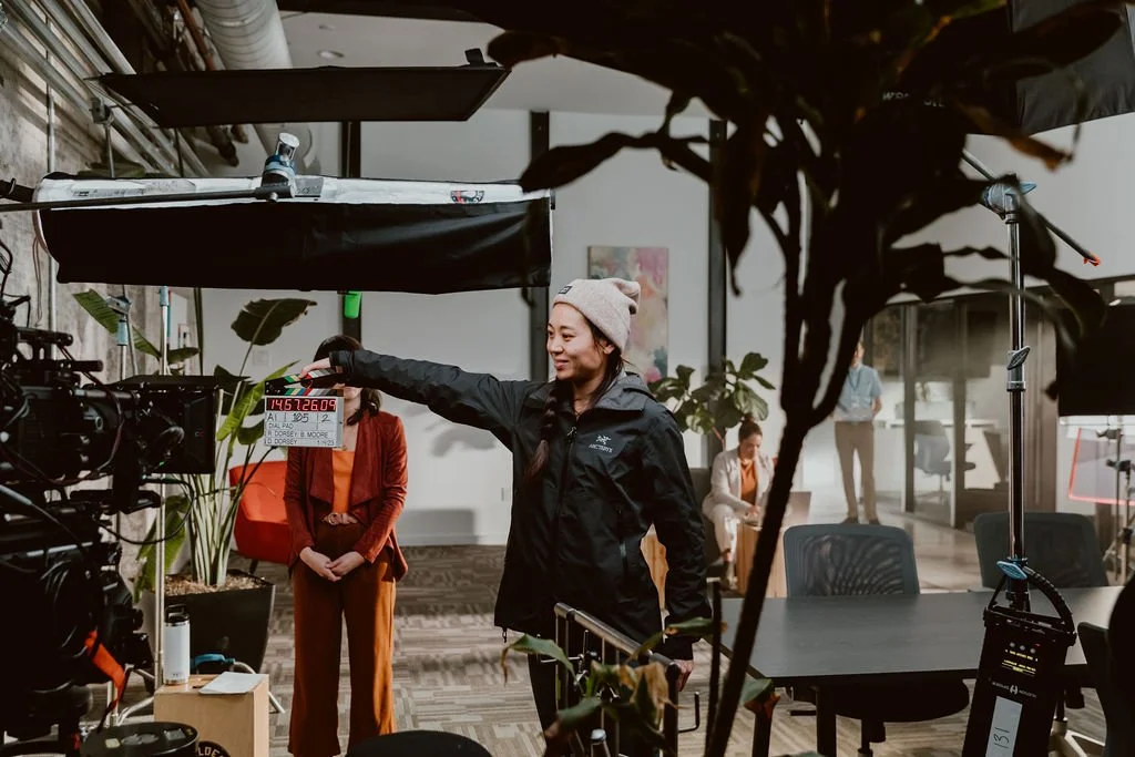 A woman on a film set holding a clapperboard, with other crew members and equipment visible in the background.