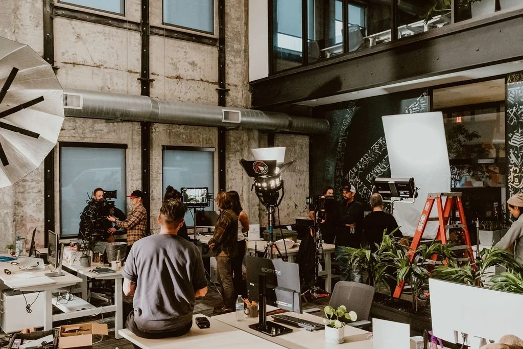 A behind-the-scenes view of a filming or photo shoot in an industrial-style office space with crew members, cameras, and lighting equipment.