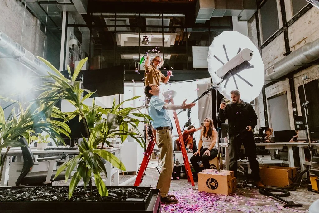 A behind-the-scenes photo of a photography or video shoot in an industrial-style office space. People are adjusting lighting equipment, including a large umbrella reflector, and a woman is standing in the background with a flower. There are plants, desks, and monitors in the foreground, with other crew members working in the background.