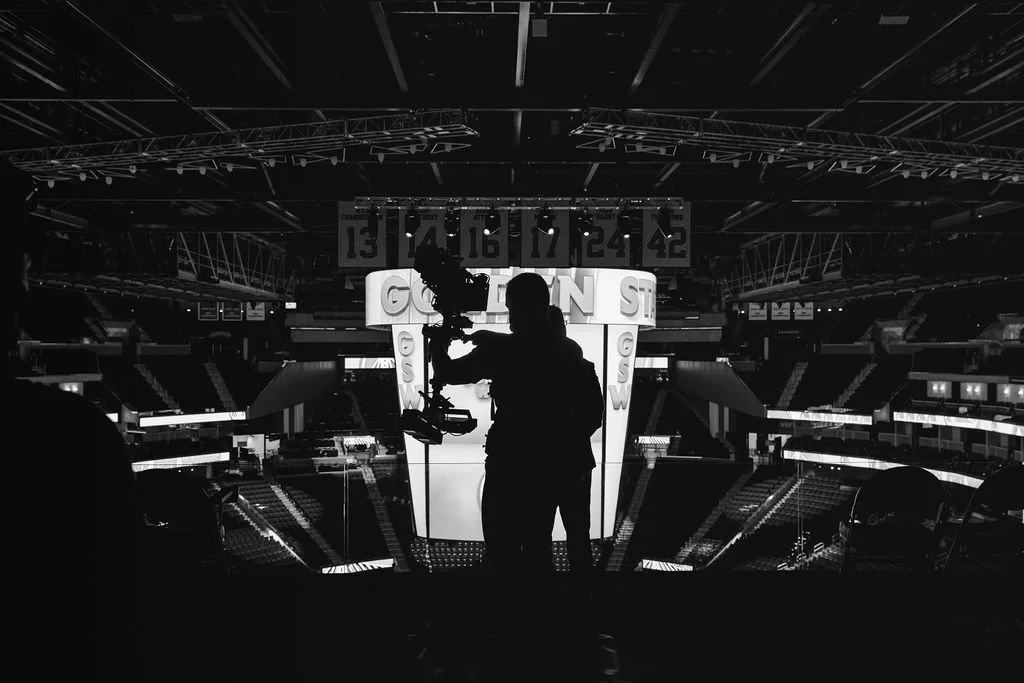 Silhouetted person filming with a camera inside an empty sports arena, with illuminated signage and scoreboard visible in the background.