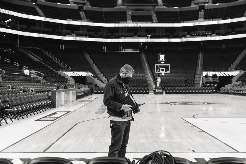 A person wearing a face mask and holding a camera and a tablet, standing alone on a basketball court inside an empty arena.