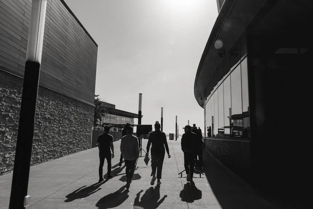 Four people walking on a sidewalk outside a modern building with glass windows, shadows cast on the ground, in a city setting.