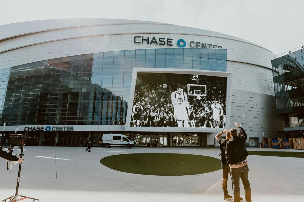 The front of Chase Center, a stadium with a large screen displaying a basketball game, with two men taking a photo in front.