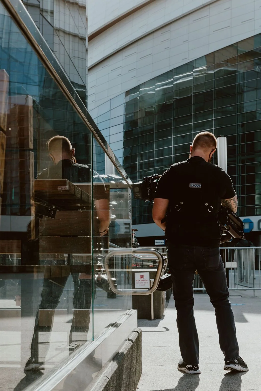 A man with a backpack and camera equipment stands outside a modern glass building, looking down and away from the camera, with his reflection visible in the glass window.