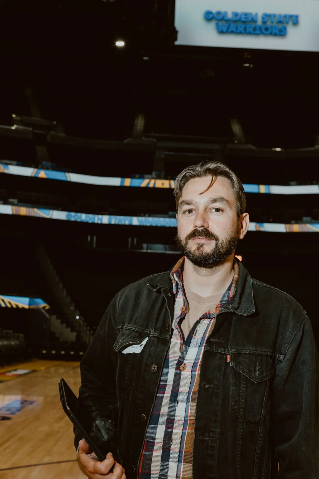 A man with a beard and styled hair stands in an indoor stadium or arena, holding a clipboard or folder. He is wearing a black denim jacket over a plaid shirt. Behind him, there is a digital screen displaying the words 'Golden State Warriors'.