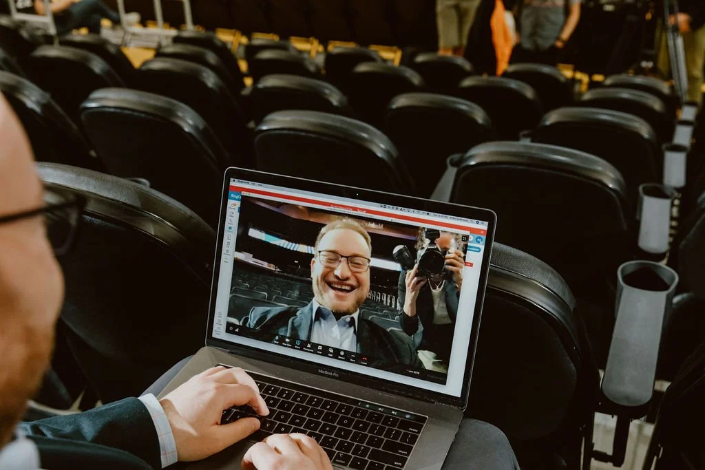 A person in glasses is taking a selfie with a laptop in a theater or conference setting, capturing their reflection on the laptop screen which shows smiling faces and a photographer.
