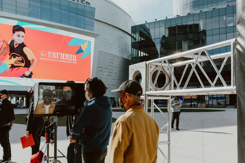 People wearing masks standing in front of a camera setup outside a modern building with a large digital billboard advertising a summer event.