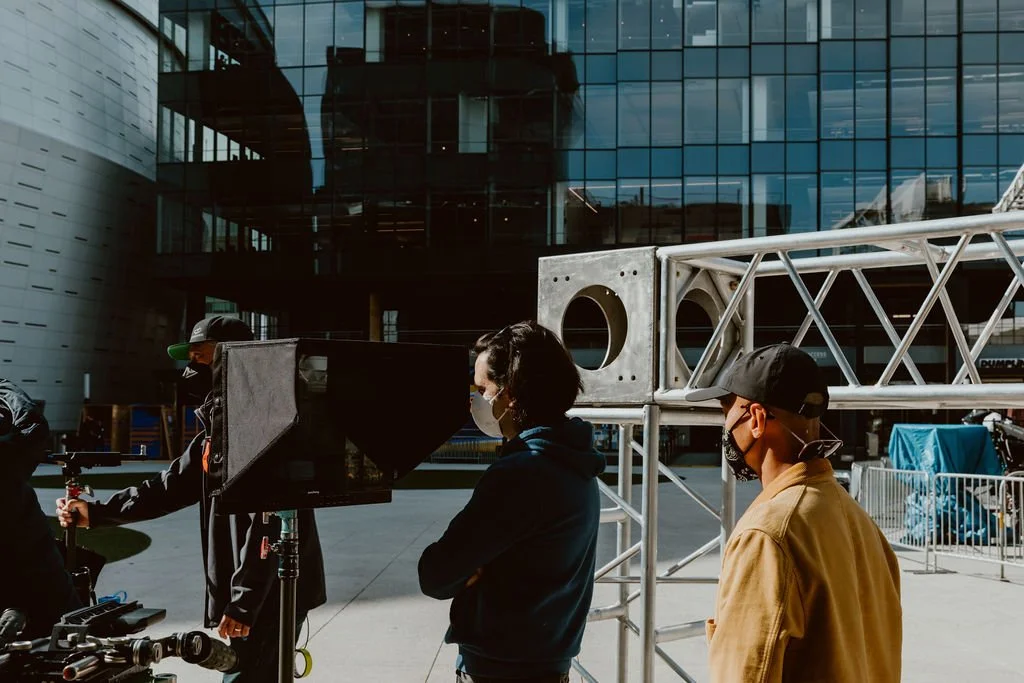 Three people wearing masks standing outdoors near a filming setup with equipment and metal scaffolding, in front of a modern glass building.