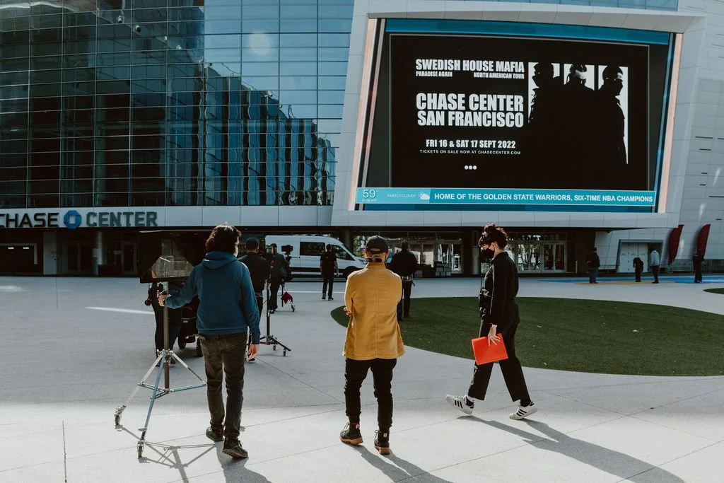 People standing outside Chase Center in San Francisco, with a large digital billboard advertising Swedish House Mafia concert and a few media crew preparing for filming.