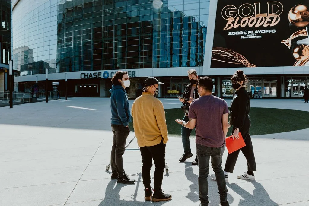 Group of five young adults standing outside in front of Chase Center, wearing masks, engaged in conversation, with a large digital billboard displaying 'GOLD BLOODED 2023 PLAYOFFS' in the background.