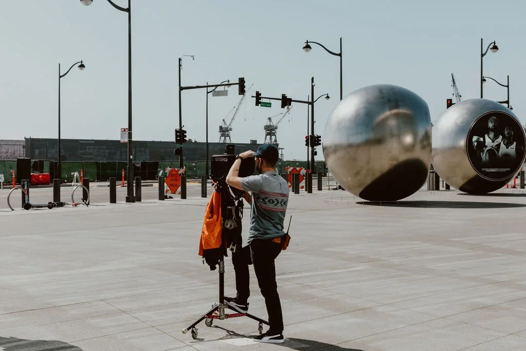 A person setting up a camera on a tripod at an outdoor urban space with large steel spherical sculptures and industrial cranes in the background.