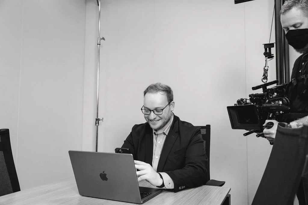 Man with glasses smiling at a laptop while another person films him with professional camera equipment in an office setting.