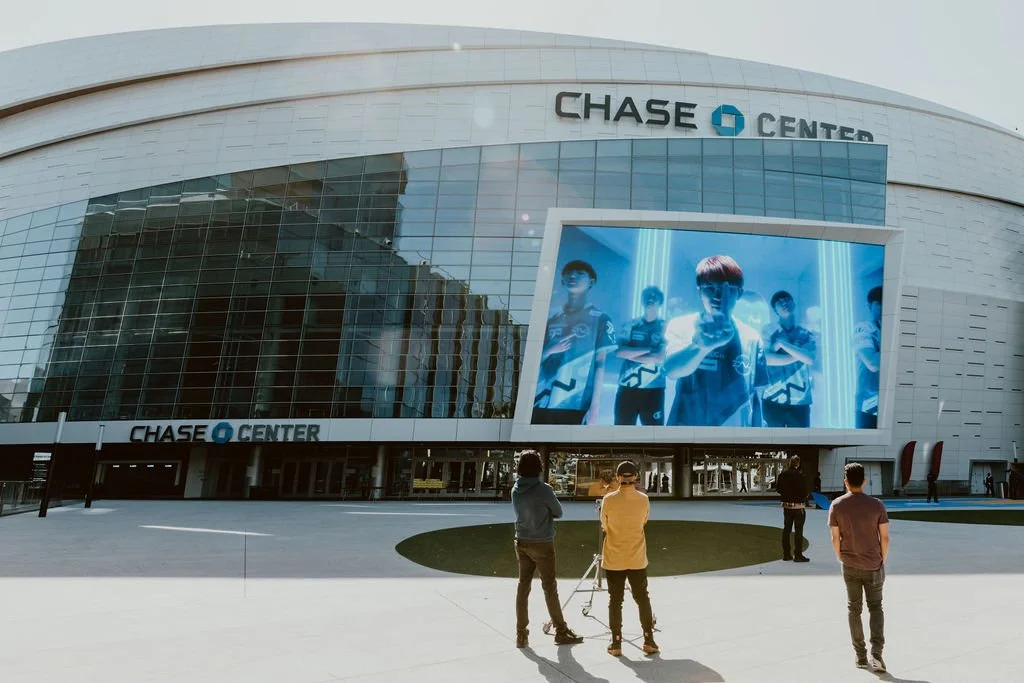 Exterior of Chase Center building with large digital screen displaying an esports team, and three people standing outside.