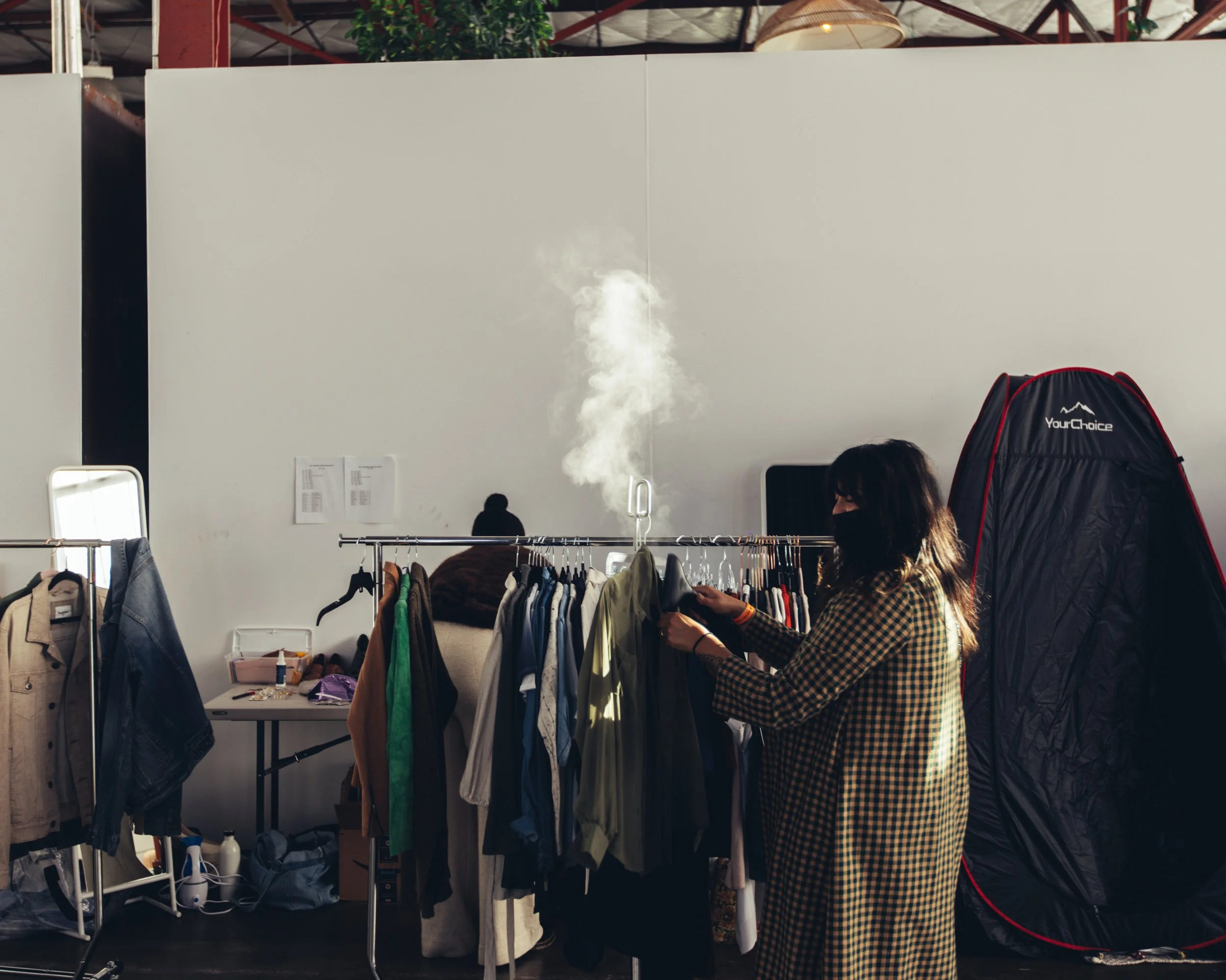A woman with dark hair wearing a plaid shirt selecting clothes from a rack at a thrift store or flea market. There are jackets and other garments hanging on the rack, with a white wall and some paperwork in the background.