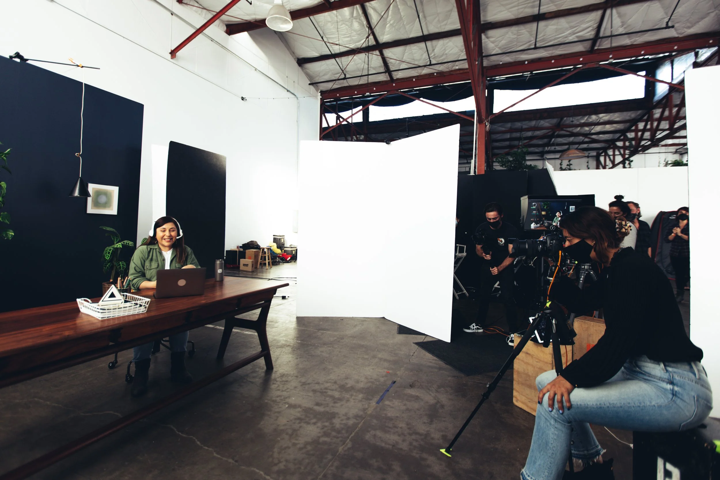 A woman in a green jacket is sitting at a desk with a laptop, smiling. The setting appears to be a studio or workspace with cameras and crew filming her. There are white and black walls, plants, and a high ceiling with exposed beams.