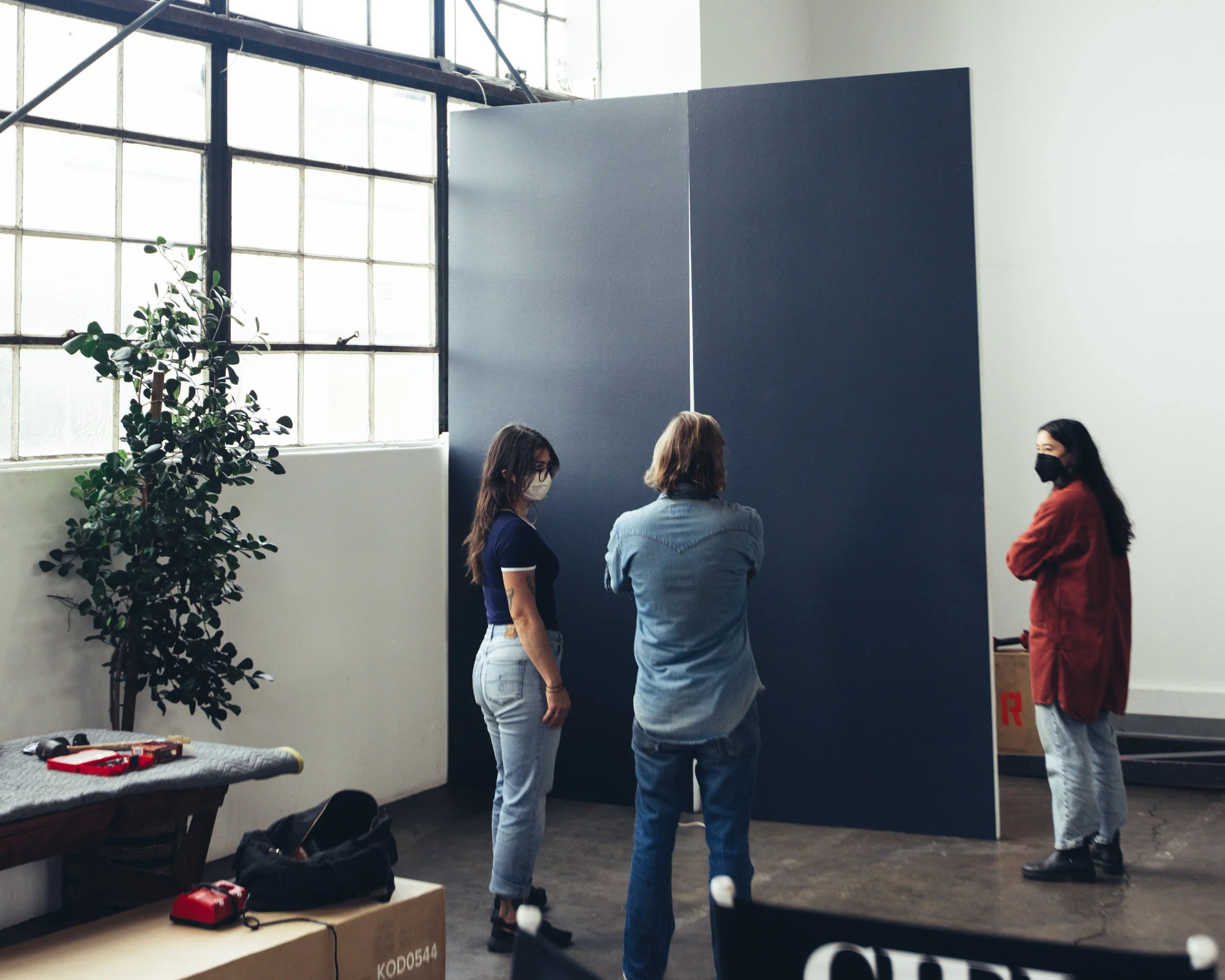 Four women wearing face masks and casual clothing are standing and talking indoors near a dark blue wall. Large windows letting in natural light are behind them, and a green potted plant is visible on the left. A table with tools and a bag is in the 