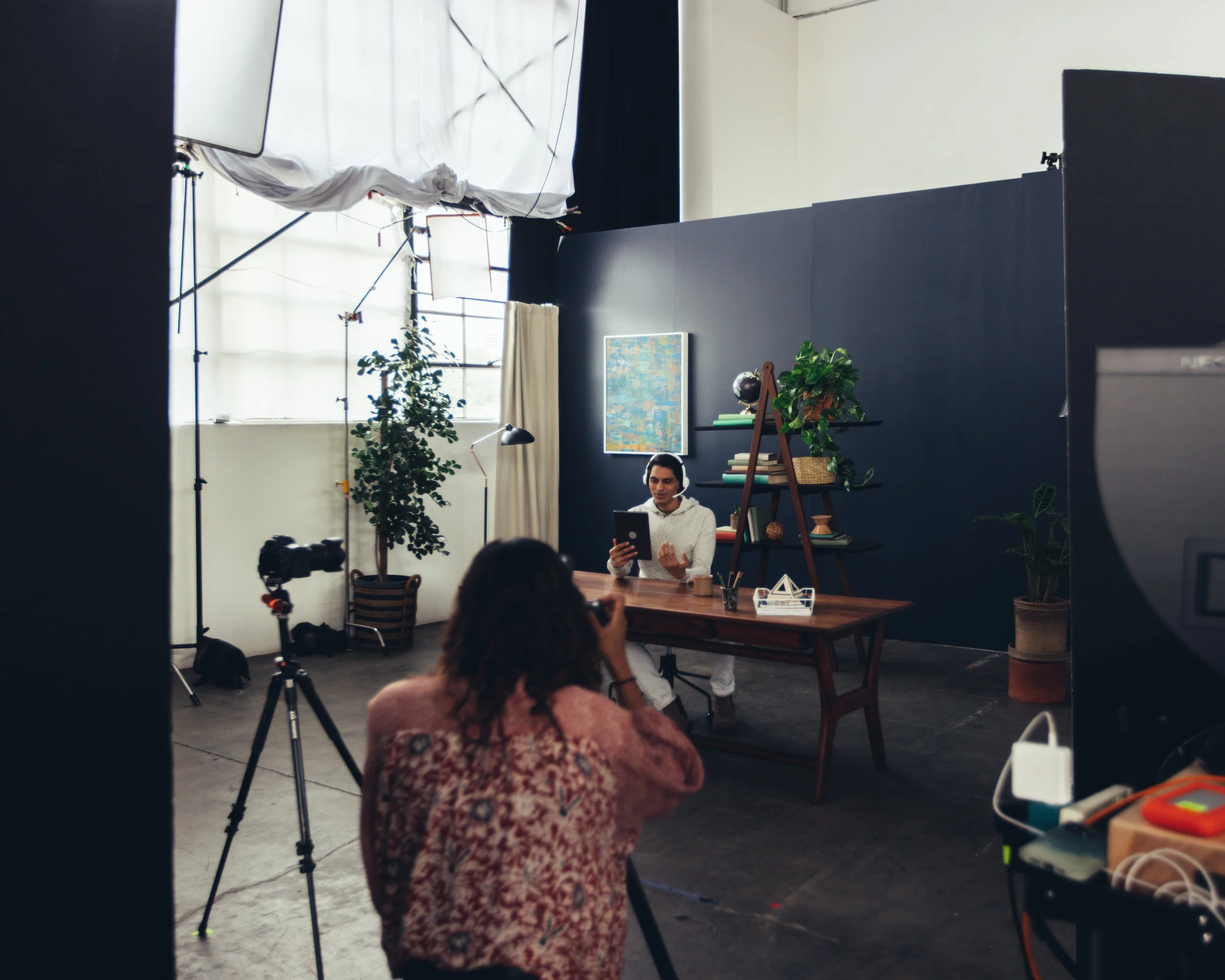 A behind-the-scenes look at a photoshoot or video shoot in a studio, with a man sitting at a table using a tablet, surrounded by lighting equipment, plants, and artwork on a dark wall.