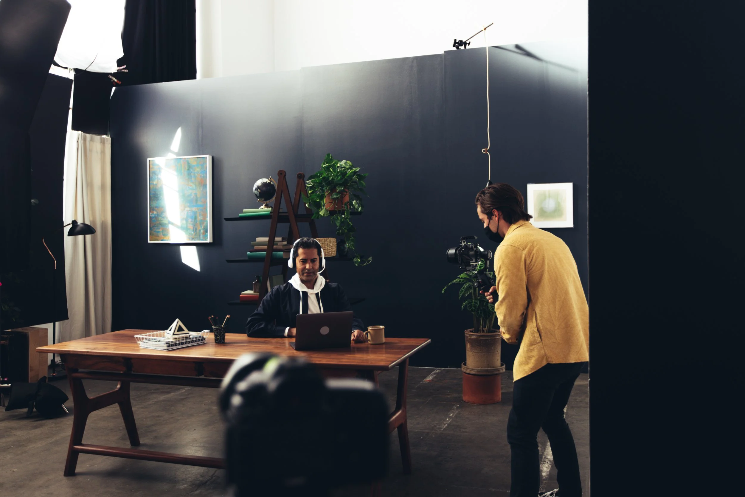 Two men in a studio, one seated at a wooden desk with a laptop and the other standing next to him holding a camera, while a third person, partially visible at the bottom, appears to be filming or observing. The background features a dark wall with fr