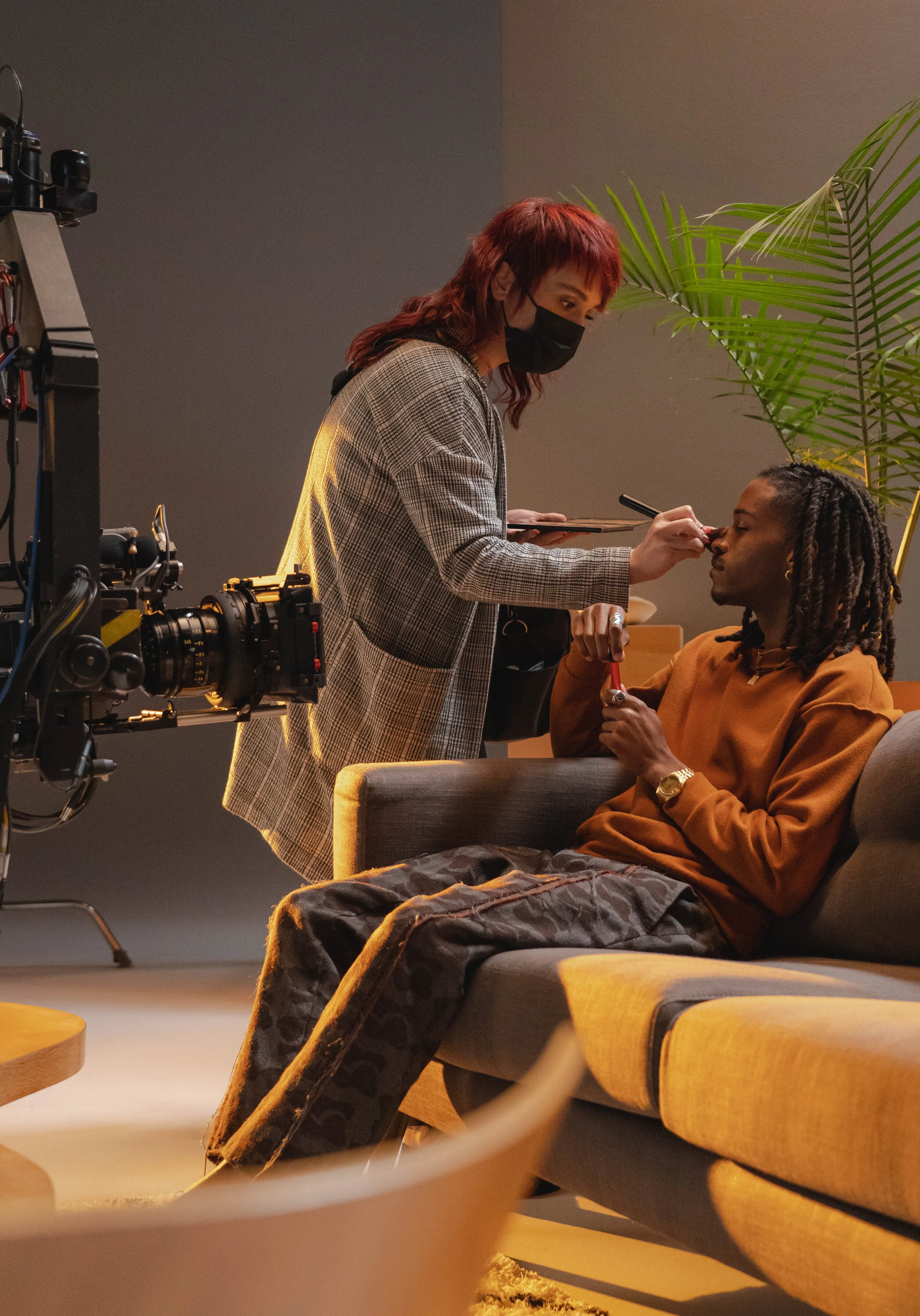 A woman with red hair and a black face mask applies makeup or tattoo ink near the eye of a man sitting on a sofa. The man is holding a small object, possibly makeup or tattoo tools. There are large green plants and a film camera in the background, an