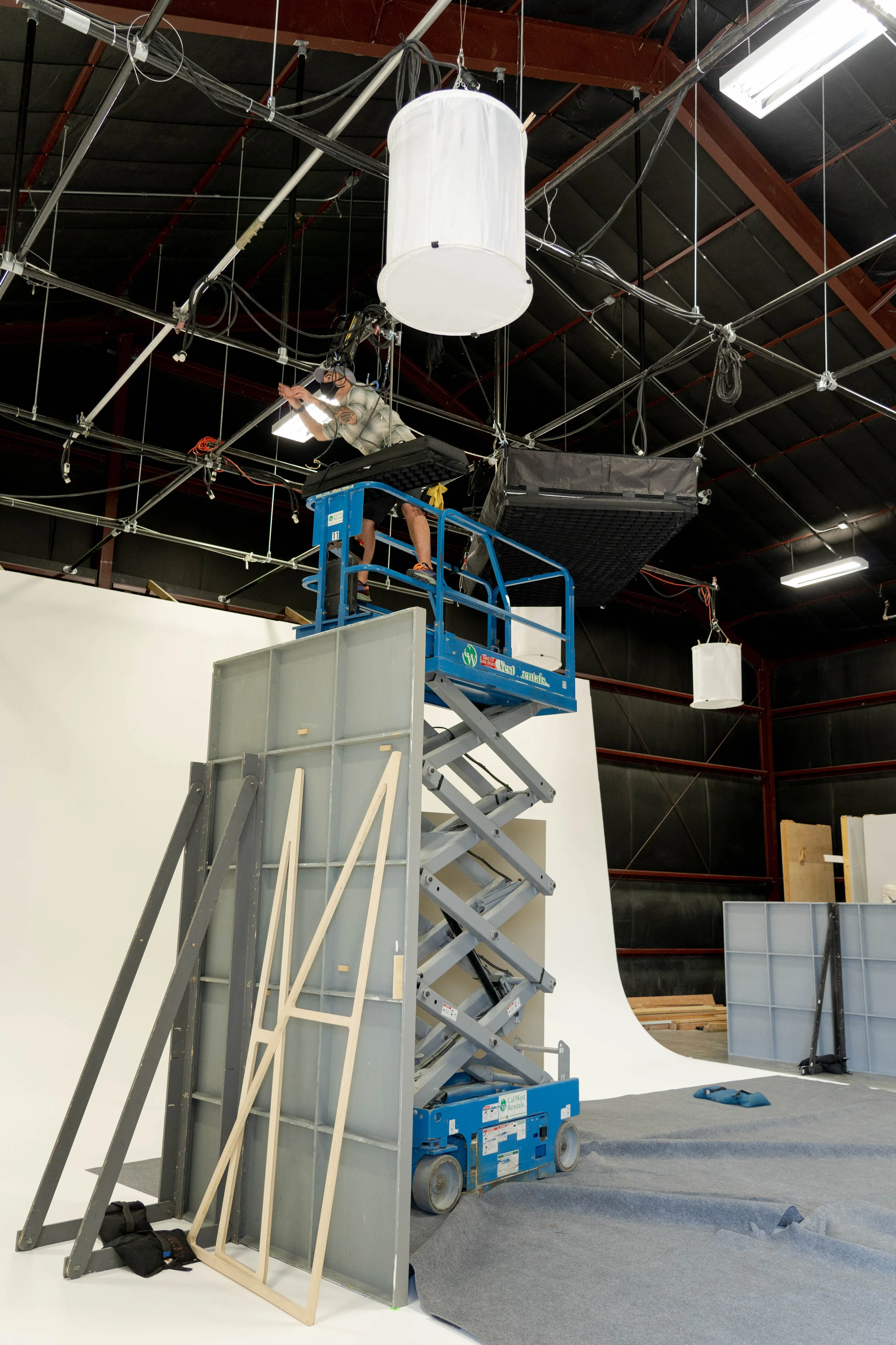 A person is working on lighting and equipment setup on a scaffolding lift in a studio with a white Cyclorama wall, some tools, and fabric on the floor.