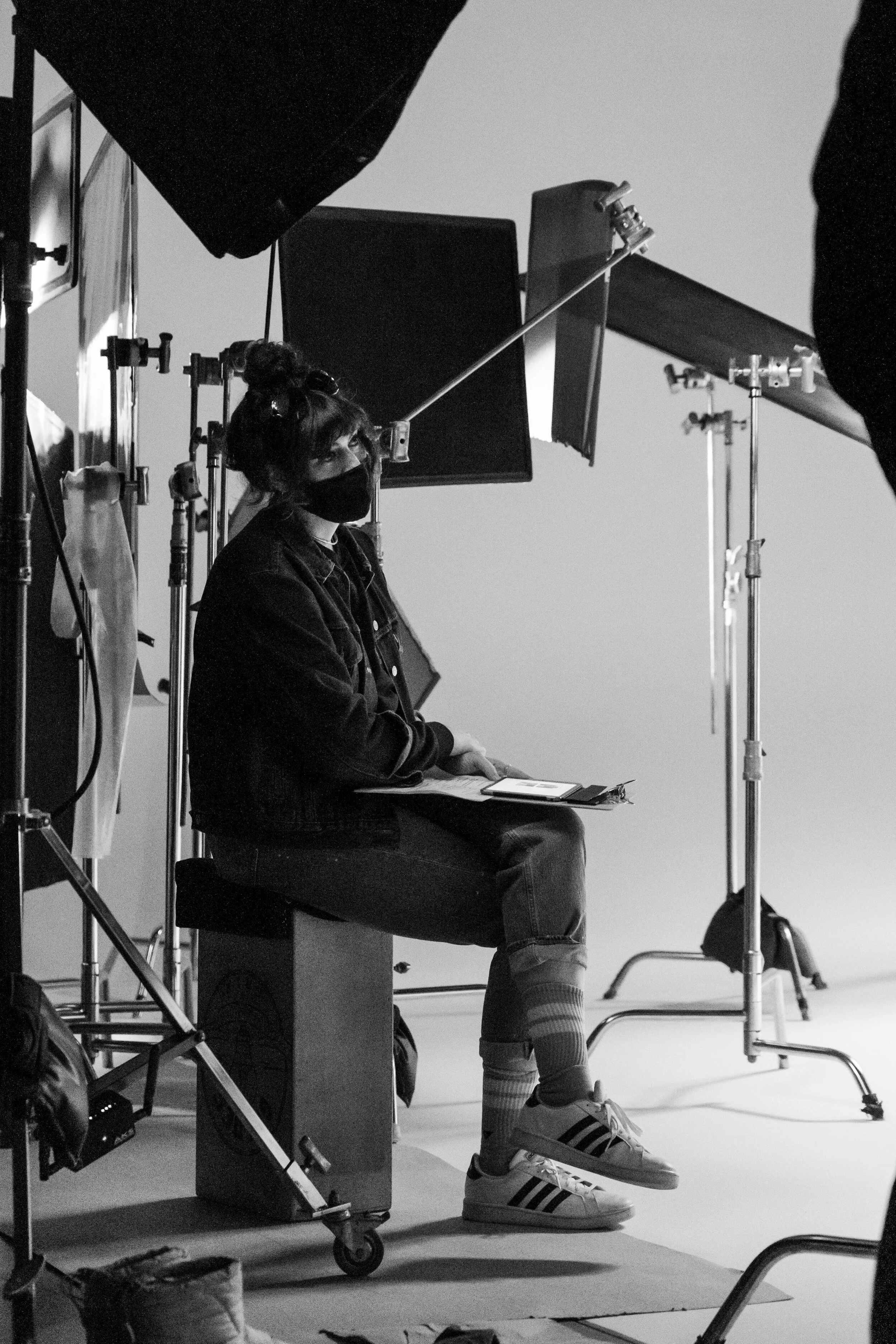 A person with a face mask and striped socks sitting on a stool in a photography studio, surrounded by lighting and photographic equipment.
