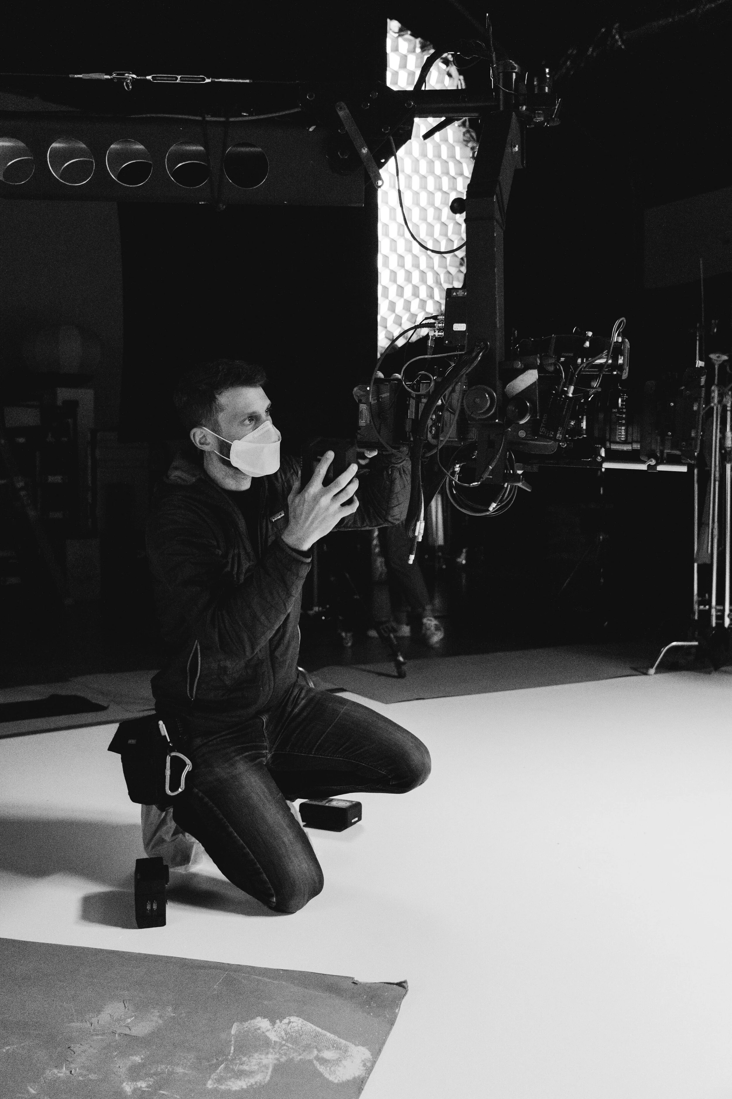 A person wearing a face mask is kneeling on one knee in a photography studio, operating a camera setup. The background includes studio lighting equipment and a textured backdrop.