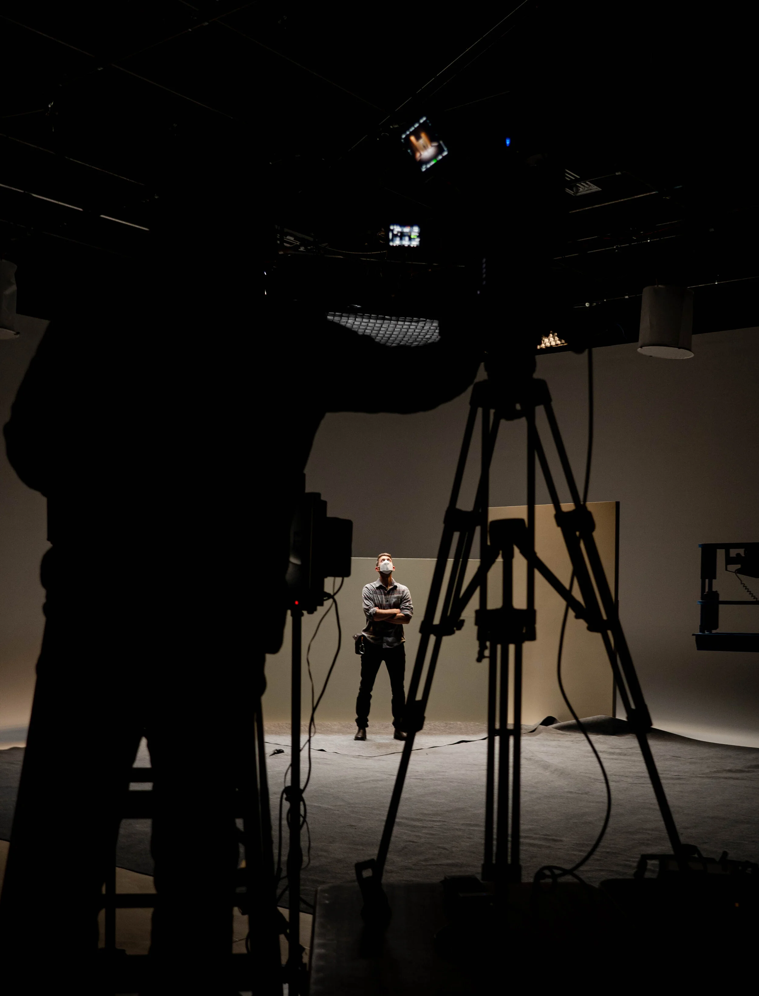 A person in the background wearing a face mask standing with arms crossed in front of a plain wall. In the foreground, there are silhouettes of camera equipment and a person working with technical gear, with part of the ceiling visible.