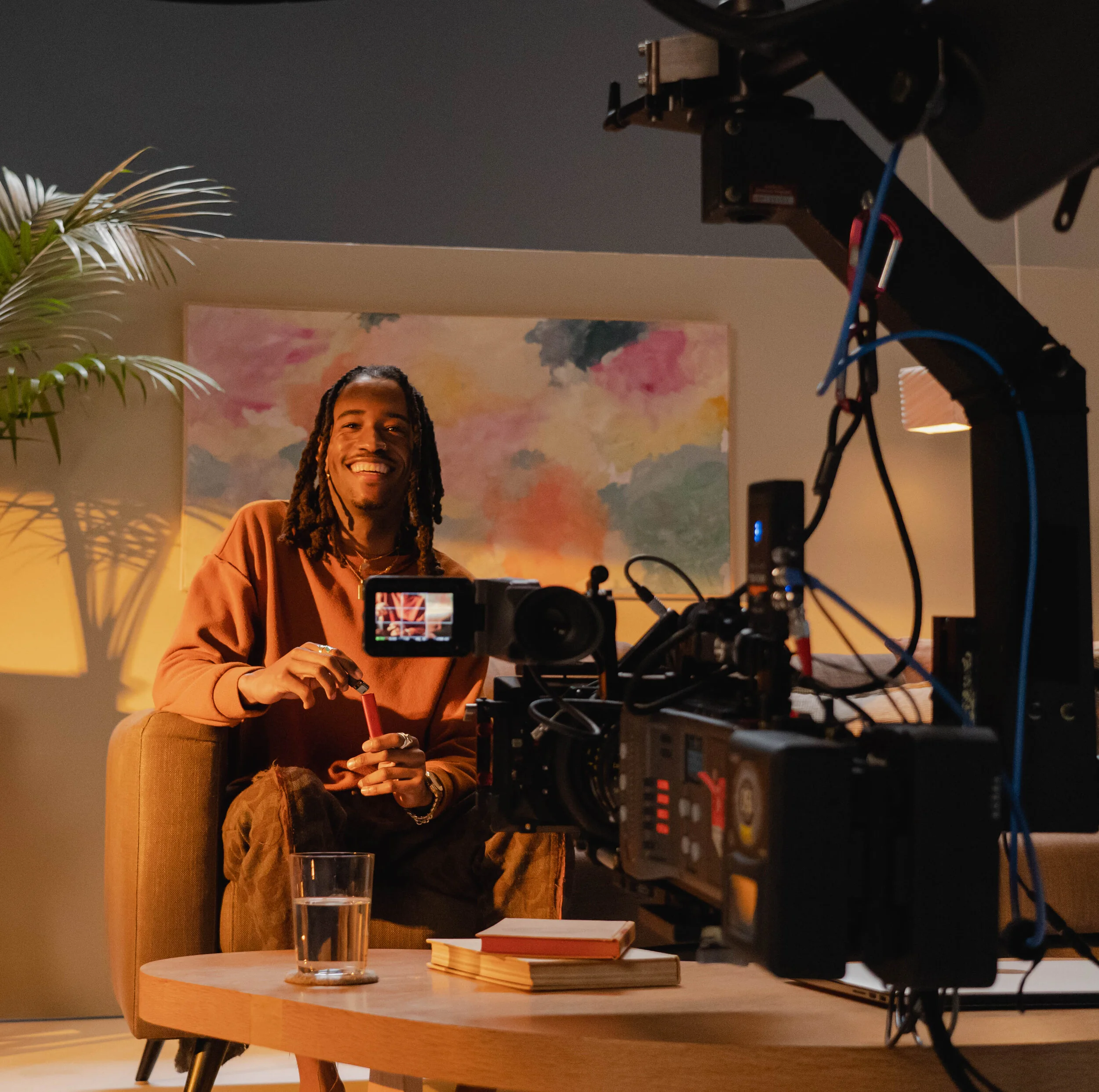 A person with dreadlocks smiles while sitting at a table with a glass of water, a closed notebook, and a closed orange book in a warmly lit room with a large colorful painting on the wall, a potted plant, and professional filming equipment in the for